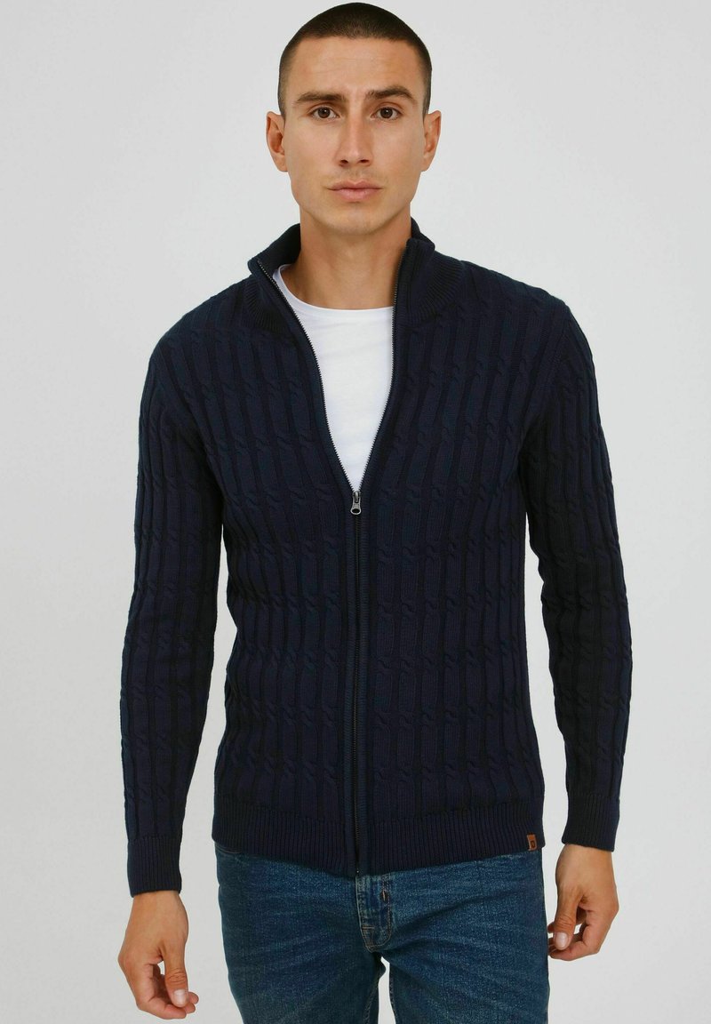 Mørkeblå strikket zip-up sweater med ribbede manchetter, der har et tekstureret kabelmønster og en høj krave. Sammen med en hvid T-shirt.