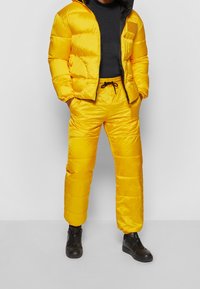 Person i en ljusgul puffjacka och matchande byxor med svarta sneakers och en svart tröja, stående mot en enkel bakgrund.