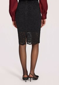 Czarna koronkowa spódnica ołówkowa z tylnym rozcięciem, z falbaniastym dołem i szczegółowymi wzorami kwiatowymi, zestawiona z czarnymi szpilkami.
