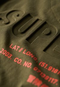 Tejido verde oliva con texto en relieve "SUPER"; los detalles impresos en rojo incluyen coordenadas geográficas y un número de empresa.