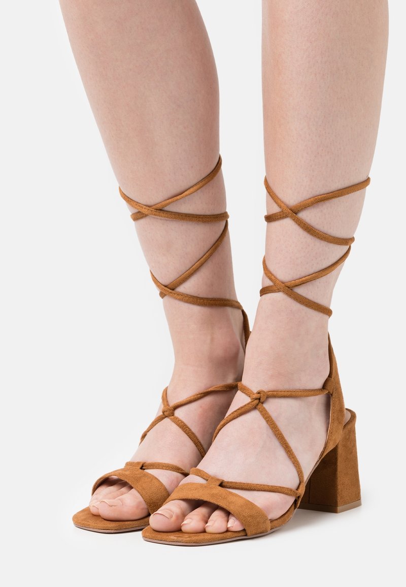 Pieds portant des sandales à talons hauts en daim marron avec plusieurs fines lanières enroulées autour des chevilles et nouées au-dessus des mollets.