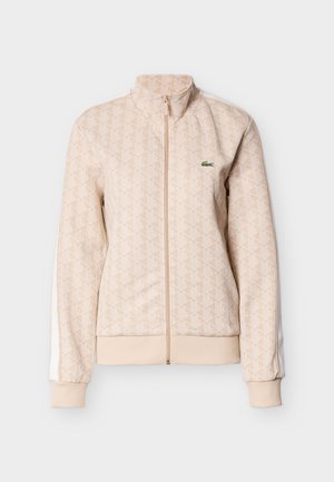Beige Jacke mit Reißverschluss und geometrischem Muster, hohem Kragen, gerippten Bündchen und Saum, weißen Seitenstreifen und kleinem grünem Krokodillogo auf der Brust.