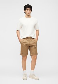 Άνδρας με σγουρά μαλλιά που φοράει λευκό πουκάμισο με κοντά μανίκια, καφέ γυρισμένα σορτς, λευκές κάλτσες και μπεζ αθλητικά παπούτσια, στέκεται με τα χέρια στις τσέπες.