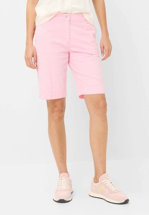 Persoon die lichtroze shorts tot op de knie, een crèmekleurig topje en bijpassende roze sneakers draagt, staand tegen een effen witte achtergrond.