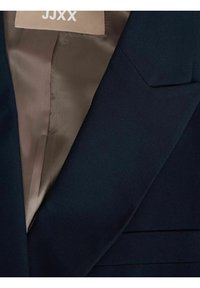 Marinblå blazermed en slät textur, med en snedkrage och ett satinfoder på insidan. Etiketten med texten "JJXX" sitter fast på insidan.