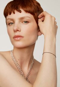 Una cadena de plata cuelga alrededor del cuello; el modelo lleva una pulsera negra delgada con un pequeño acento dorado en la muñeca, mostrando un tono de piel suave.