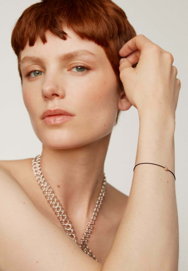 Una cadena de plata cuelga alrededor del cuello; el modelo lleva una pulsera negra delgada con un pequeño acento dorado en la muñeca, mostrando un tono de piel suave.