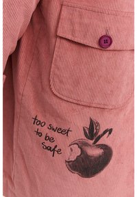 Lososová ružová kordová látka s vertikálnymi ryhami. Obsahuje klopovú kapsu s fialovým gombíkom a čiernu grafiku jablka s textom.