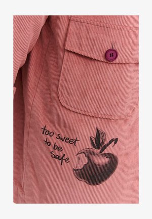 Lososově růžová kordurová látka s vertikálními žebry. Obsahuje klopovou kapsu s fialovým knoflíkem a černou grafikou jablka s textem.