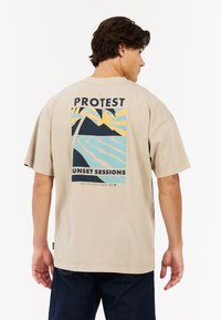 Beige t-shirt met korte mouwen, voorzien van een grafisch ontwerp op de achterkant met bergen, een zon en golven. De tekst luidt "PROTEST" en "SUNSET SESSIONS."