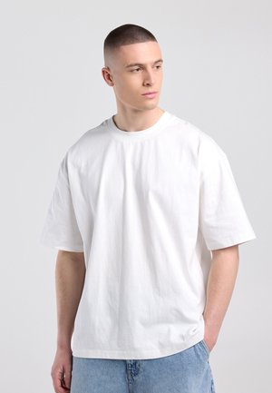 Jonge man met kort haar die een effen wit oversized T-shirt en lichtblauwe spijkerbroek draagt, kijkt naar links tegen een grijze achtergrond.