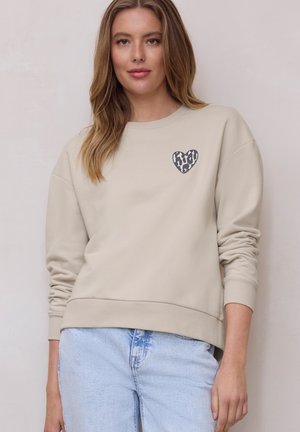 Femme aux longs cheveux châtain clair portant un sweat-shirt beige avec un cœur imprimé léopard sur la poitrine et un jean bleu clair.