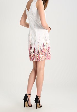 Mujer con vestido blanco sin mangas con estampado floral rosa en el borde, calzando zapatos negros de tacón alto con correa en el tobillo, de pie con la espalda hacia la cámara.