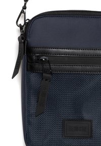 Námořnická crossbody taška vyrobená z texturované tkaniny s hladkým černým zipem, obsahující značenou nášivku a nastavitelné ucho pro všestranné použití.