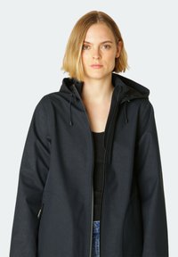 Chaqueta ligera y oscura con capucha, con cierre de cremallera en el frente, tela texturizada y cordones ajustables. Sin patrones o acentos prominentes.