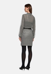 Schwarz-weiß gemustertes Kleid mit transparenten langen Ärmeln, tailliertem Gürtel und eng anliegendem Rock. Getragen mit schwarzen Strumpfhosen und spitzen Absätzen.