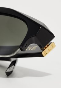 Lunettes de soleil noires avec une monture brillante, des verres verts et des accents en métal doré, présentant un détail de marque en or sur le côté avec "RSF" en relief.