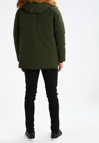 Grüne Parkajacke mit einem pelzgefütterten Kapuzen, die einen geraden Schnitt und eine glatte Textur aufweist. Getragen über schwarzen Skinny-Jeans und Turnschuhen.