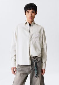 Light beige button-up shirt with a patch pocket, paired with faded gray jeans featuring a carabiner and keychain accessory. Smooth fabric.