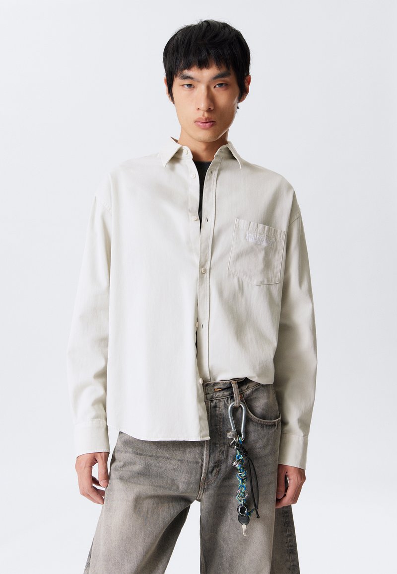 Light beige button-up shirt with a patch pocket, paired with faded gray jeans featuring a carabiner and keychain accessory. Smooth fabric.