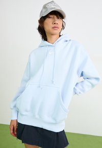 Hoodie azul claro com um bolso frontal, cordão ajustável e logótipo branco da Nike. Combinado com uma saia preta e um boné cinza.