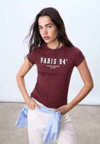 Burgundská bavlněná trička s potiskem 'PARIS 94' a 'CONFIDENCE CLUB' v bílé barvě. V kombinaci s bílými kalhotami a modrou pruhovanou košilí uvázanou v pase.