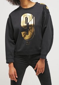 Sweat-shirt noir avec un grand graphique doré "9" sur le devant, doté d'un col rond et d'une coupe ample. Comprend des accents aux épaules.