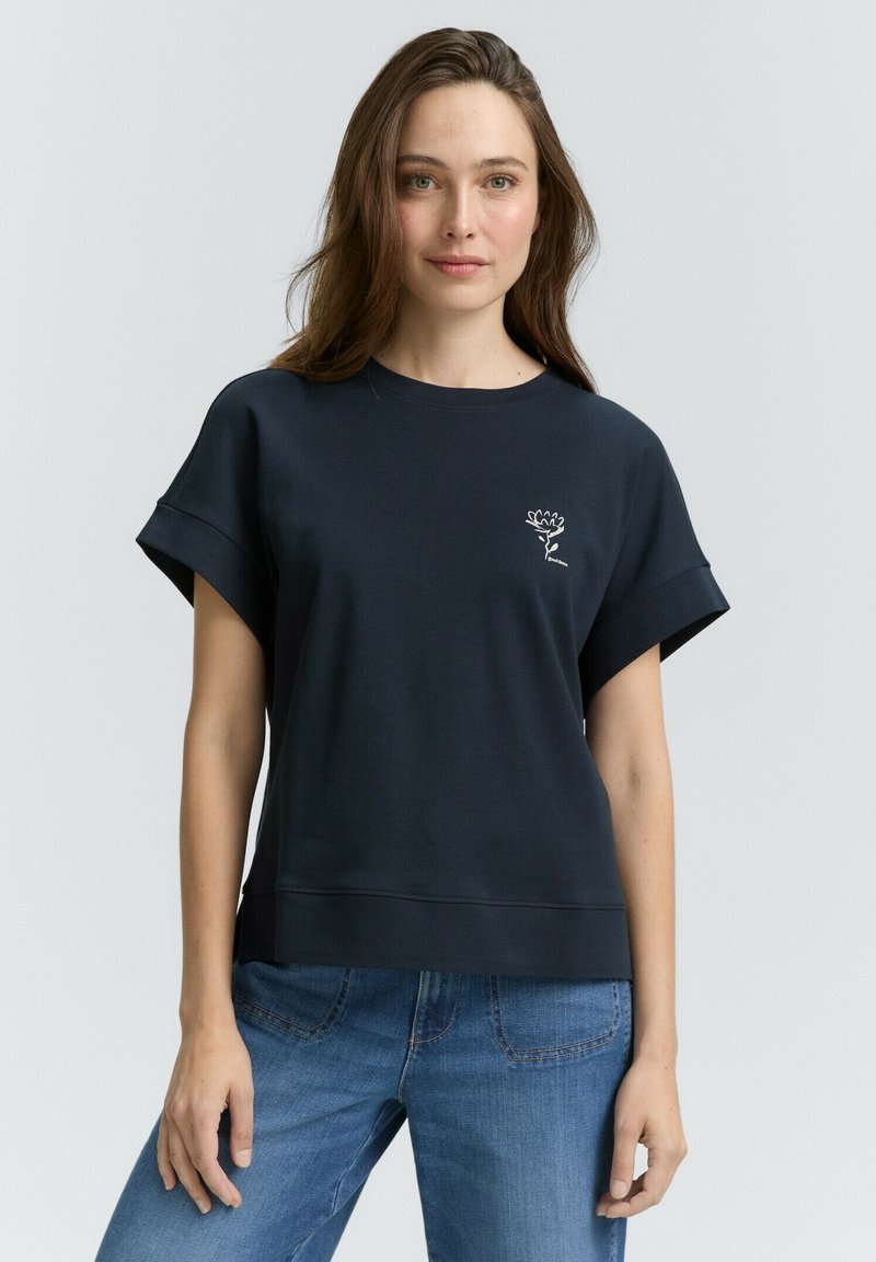 Sweatshirt en coton bleu marine à manches courtes et à ourlet côtelé, arborant un graphique floral blanc sur la poitrine. Porté avec un jean bleu clair.