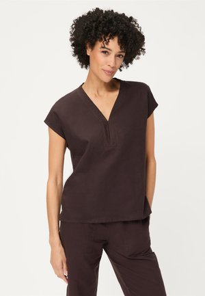 Donna con capelli ricci che indossa una maglia marrone scuro a maniche corte con scollo a V e pantaloni abbinati su sfondo chiaro e semplice.