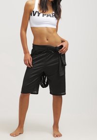 Donna che indossa un top corto bianco con scritte e pantaloni shorts neri in rete larghi, in piedi a piedi nudi su uno sfondo semplice.