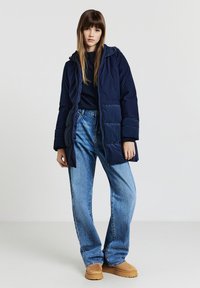 Giacca imbottita blu navy con cappuccio, dotata di chiusura con zip, tasche laterali e pannelli testurizzati a contrasto. Abbinata a jeans a gamba larga azzurri.