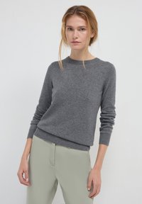 Jeune femme aux cheveux blonds portant un pull gris à manches longues et un pantalon gris clair, se tenant devant un fond blanc uni.