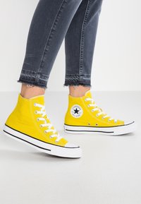 Gule sneakers med højt skaft i lærred, med hvide snørebånd, sorte detaljer og et rundt logo på siden. Bruges med mørke, opslidte jeans.