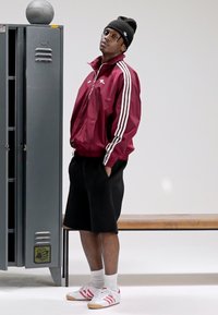 Corta-vento bordô com riscas brancas, calções pretos e ténis brancos com riscas vermelhas. Em pé ao lado de um cacifo cinzento, com uma bola de basquetebol por perto.