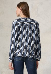 Blusa a maniche lunghe in un motivo astratto blu navy, azzurro e bianco. Presenta un giromanica con bottoni e un tessuto morbido. Abbinata a jeans blu scuro.