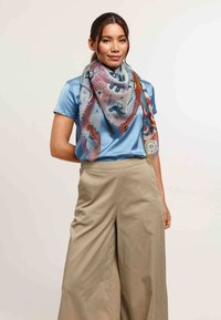 Pañuelo de seda con un diseño colorido que presenta tonos de rosa, azul y naranja, lucido elegantemente alrededor del cuello, combinado con una blusa azul y pantalones beige.