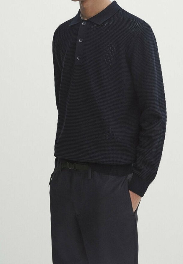 Homme portant un polo noir texturé à manches longues rentré dans un pantalon noir, debout avec une main dans la poche.