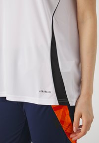 Witte sportshirt met zwarte zijpanelen en "AEROREADY" logo. Korte mouwen, gladde textuur, gedragen met marineblauwe broek met een oranje geometrisch patroon.