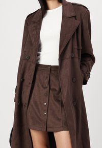 Czarny zamszowy płaszcz trench z podwójnym zapięciem, w połączeniu z pasującą brązową spódnicą z frontowymi zatrzaskami i gładką teksturą.