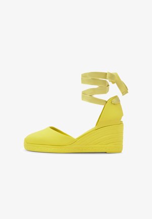 Gelber Espadrille-Keilabsatzschuh mit geschlossenem Zehenbereich, strukturiertem Absatz und langen Stoffbändern, die um den Knöchel gewickelt sind.