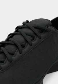 Κοντινό πλάνο μαύρου δερμάτινου αθλητικού παπουτσιού με υφή και σφιχτά δεμένα επίπεδα μαύρα κορδόνια σε λευκό φόντο.