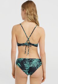 Donna con lunghi capelli biondi che indossa un bikini nero con motivo a foglie di palma verdi, vista di spalle su uno sfondo bianco uniforme.