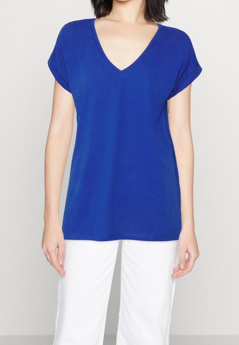 Mujer con una blusa azul real de manga corta y escote en V, y pantalones blancos, de pie contra un fondo liso gris claro.