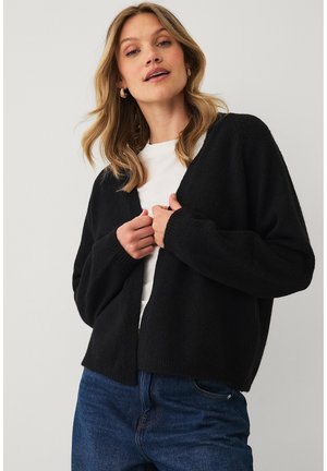 Ung kvinde iført en sort cardigan, hvid skjorte og blå jeans, der holder i cardiganens forside og kigger lidt opad.