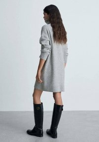 Robe en laine grise avec un col haut, des manches longues et des poignets côtelés. Associée à des bottes en cuir noires montantes avec une boucle dorée.