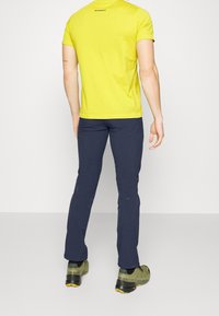 T-shirt jaune à manches courtes avec logo, associé à un pantalon bleu foncé. La chaussure est verte avec des accents noirs et jaunes. Fond neutre.
