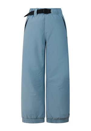 Jongen draagt een zwarte beanie, een tweekleurige blauw-beige longsleeve, losse blauwe broek met riem en zwarte laarzen, staand met handen in de zakken.