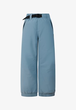 Jongen draagt een zwarte beanie, een tweekleurige blauw-beige longsleeve, losse blauwe broek met riem en zwarte laarzen, staand met handen in de zakken.