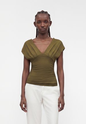 Donna con capelli intrecciati che indossa un top verde oliva a scollo a V con dettagli raccolti e pantaloni bianchi, in piedi davanti a uno sfondo chiaro e uniforme.