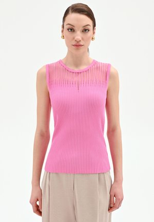 Donna con capelli castani, indossa un top rosa senza maniche a coste con scollo trasparente, pantaloni beige e orecchini a cerchio dorati, in piedi davanti a uno sfondo bianco.